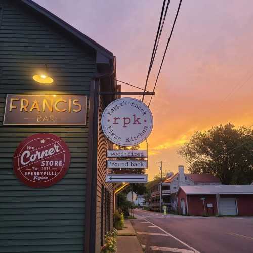 A colorful sunset behind a building with signs for "Francis Bar" and "rpk Pizza Kitchen" on a quiet street.