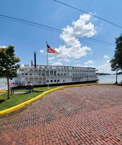 A large steamboat docked by a river, with an American flag flying and a sunny sky above. Brick pathway in the foreground.