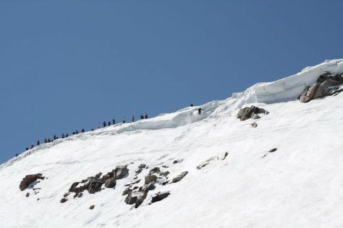 A snowy mountain peak with a line of climbers along the ridge against a clear blue sky.