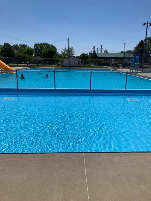 A clear blue swimming pool with a slide, surrounded by a fence and a sunny sky, with a few people swimming.