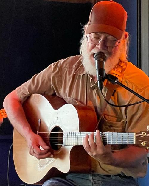 A musician with a beard and glasses plays an acoustic guitar while singing into a microphone.