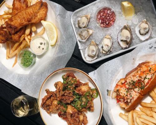 A spread of seafood dishes: fried fish and chips, oysters on ice, fried chicken with jalapeños, and a lobster roll with fries.
