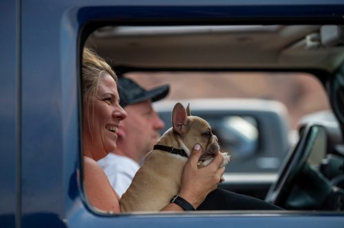 A woman smiles while holding a small dog inside a vehicle, with a man seated beside her.