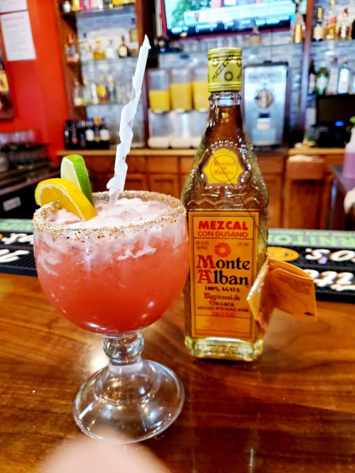 A colorful cocktail with a salted rim, garnished with lime and orange, next to a bottle of Monte Alban mezcal.