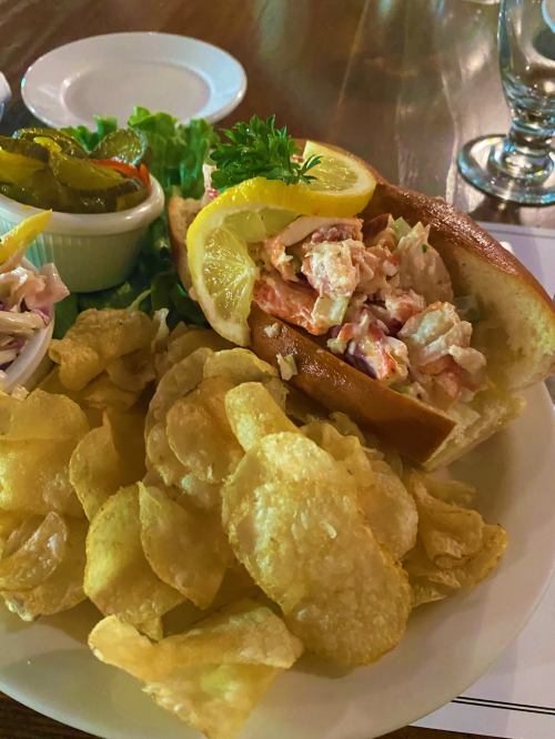 A lobster roll with lemon and lettuce, served with potato chips and a side of pickles and coleslaw.