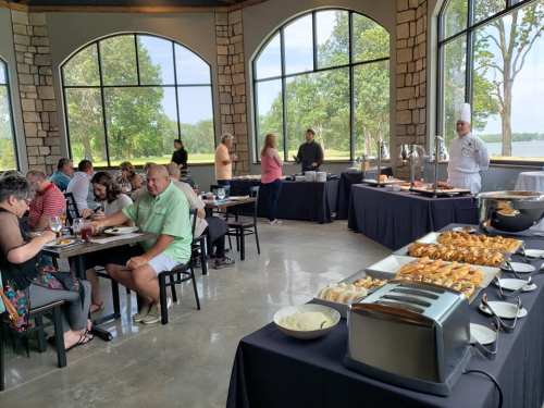 A dining area with guests enjoying a buffet, featuring a chef, tables, and large windows overlooking nature.