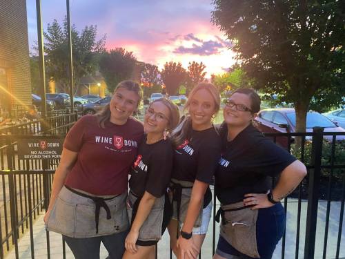Four women in matching shirts pose together outdoors at sunset, smiling in front of a restaurant.