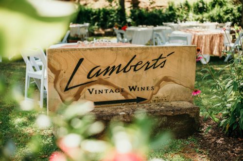Sign for "Lambert's Vintage Wines" with an arrow, surrounded by greenery and outdoor event setup in the background.