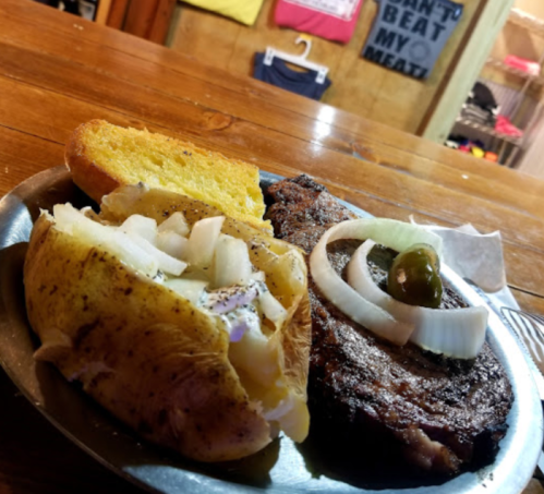 A plate with a grilled steak, baked potato topped with onions, and a slice of cornbread.