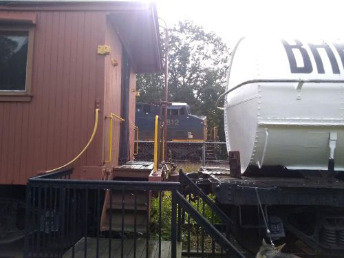 A train car next to a building, with a tank and a locomotive visible in the background under a bright sky.