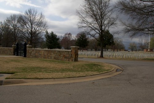A stone wall and gate lead into a cemetery with rows of white headstones under a cloudy sky.