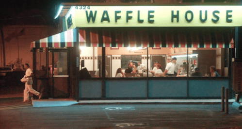 A Waffle House at night, with patrons inside and a person walking by outside. Bright yellow sign and colorful awning.