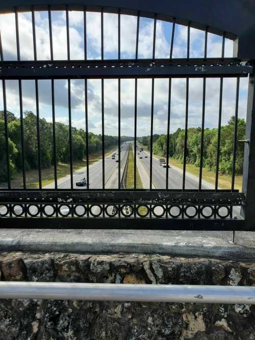 View of a highway through a metal fence, flanked by trees under a partly cloudy sky.