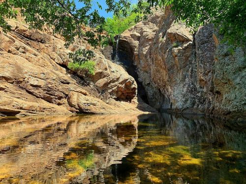A serene rocky landscape with a clear pool of water reflecting the surrounding cliffs and greenery.