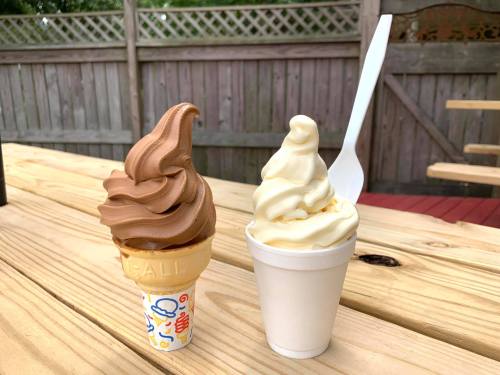 Two soft serve ice cream cones: one chocolate in a cone and one vanilla in a cup, placed on a wooden table.