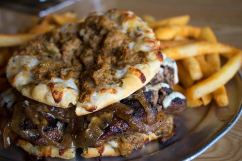 A close-up of a stacked burger with grilled onions and sauce, served with crispy French fries on a metal plate.