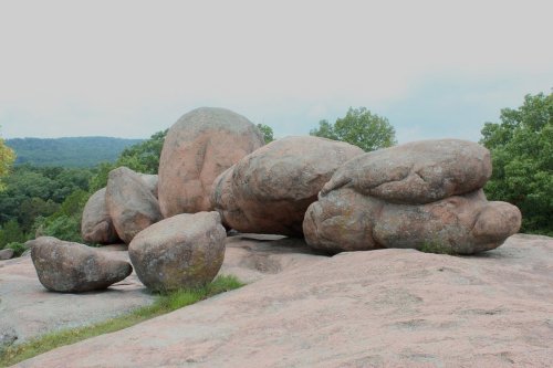 A cluster of large, smooth boulders on a rocky surface, surrounded by greenery and distant hills under a cloudy sky.