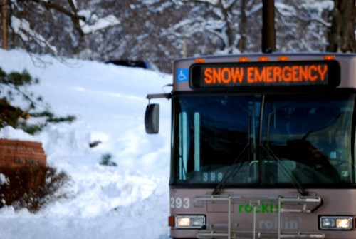 A bus with a sign reading "SNOW EMERGENCY" parked in a snowy area, surrounded by snow-covered trees.