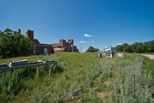 Abandoned building in the background with overgrown grass and discarded machinery in the foreground under a clear blue sky.