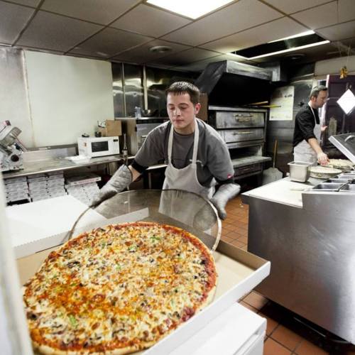 A chef prepares a large pizza in a busy kitchen, with another chef working in the background.