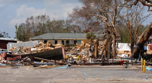 Devastated landscape showing a destroyed building and debris scattered among trees after a natural disaster.
