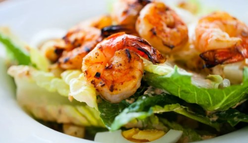 Grilled shrimp atop a fresh salad with romaine lettuce and other vegetables, served in a white bowl.