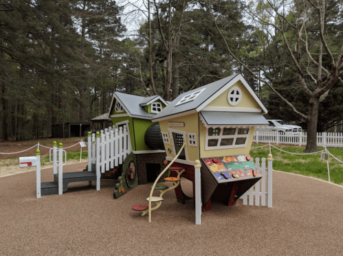 Colorful playground structure resembling houses, with slides and climbing features, surrounded by trees and a fence.