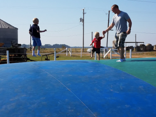 A man holds hands with a child while another child jumps on a large inflatable surface in a rural setting.