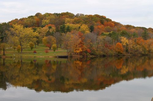 A serene lake reflecting vibrant autumn foliage on a hillside, with trees in shades of orange, yellow, and green.