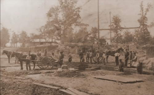 Historic black-and-white photo of workers and horses at a construction site, with trees and buildings in the background.