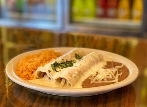 A plate of enchiladas topped with creamy sauce, served with rice and refried beans, garnished with fresh herbs.