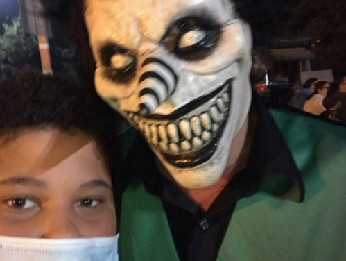 A child in a mask poses for a selfie with a person in a creepy clown mask and green vest at a nighttime event.