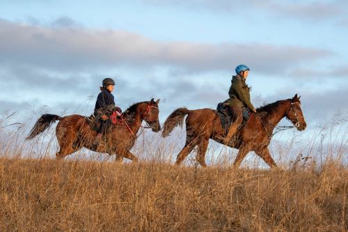 Two riders on horseback walk along a grassy hill under a cloudy sky during sunset.