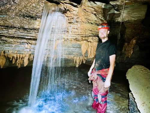 A person stands in a cave near a waterfall, surrounded by rock formations and water, wearing a headlamp and muddy gear.