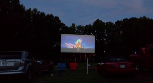 A movie screen displaying a colorful scene, surrounded by parked cars and camping chairs in a twilight setting.