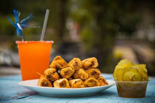 A plate of golden spring rolls, a cup of pickles, and a colorful drink with a straw on a rustic table.