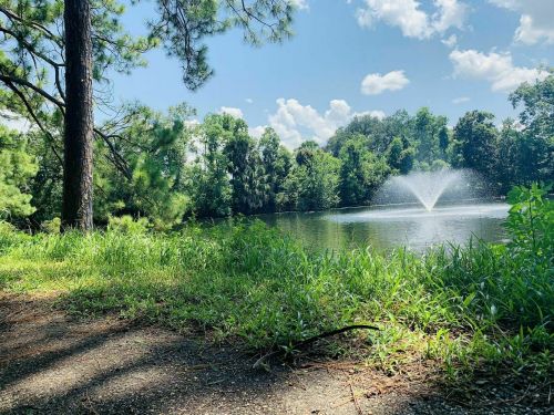 A serene pond surrounded by lush greenery, featuring a fountain spraying water under a clear blue sky.