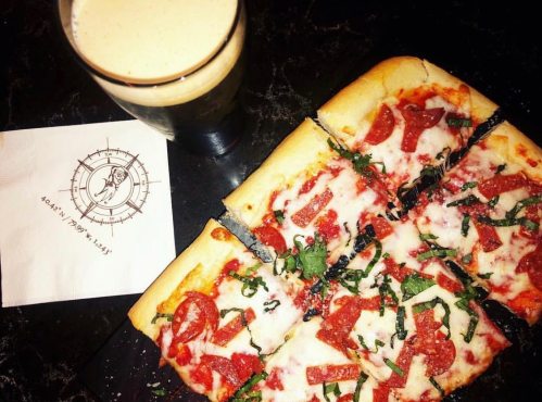 A square pizza topped with pepperoni and greens, alongside a glass of dark beer and a napkin on a black surface.