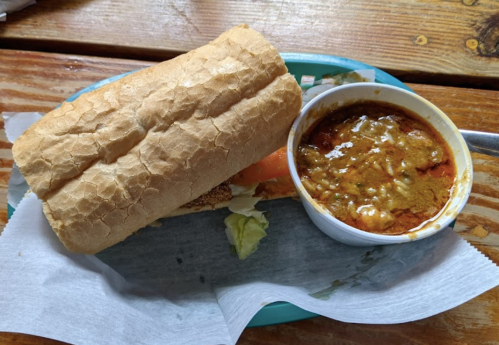 A sandwich on a plate next to a bowl of curry with cheese, served on a wooden table.