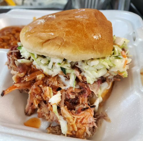 A pulled pork sandwich topped with coleslaw on a soft bun, served in a takeout container with baked beans on the side.