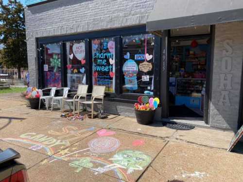 A colorful storefront with a chalk drawing on the sidewalk, featuring chairs outside and various decorations in the windows.