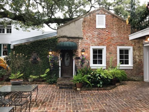 Charming brick house with green awning, surrounded by plants and a brick patio, under a canopy of trees.