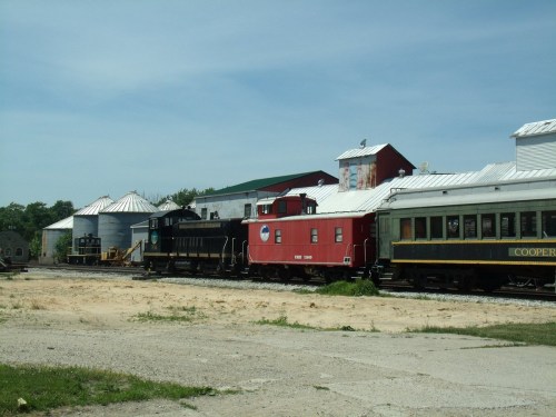 A black locomotive and a red caboose parked beside vintage train cars near a barn under a clear blue sky.