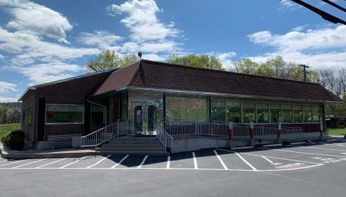 A modern brick building with large windows, surrounded by green trees and a clear blue sky. Parking spaces in front.