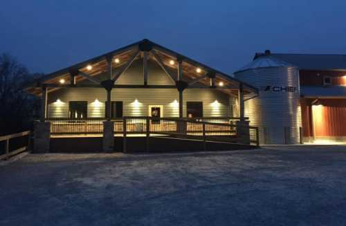 A modern barn with a lit porch and a silos, set against a twilight sky.