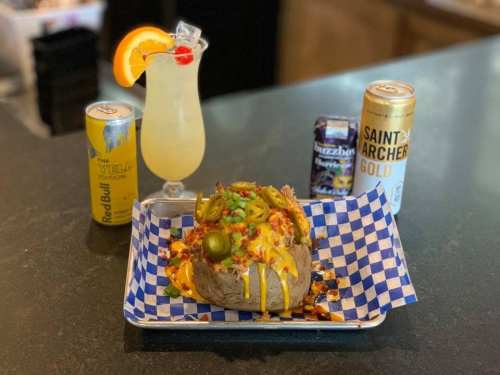 A loaded baked potato topped with cheese, jalapeños, and green onions, accompanied by drinks on a checkered plate.