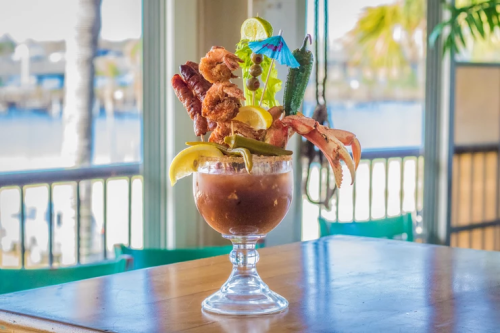A large, colorful cocktail garnished with shrimp, bacon, and vegetables, set against a waterfront backdrop.