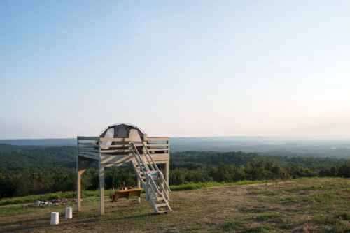 A wooden structure on stilts overlooks a vast green landscape under a clear sky.