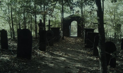 A dimly lit graveyard with old tombstones, surrounded by trees and a wrought iron gate in the background.