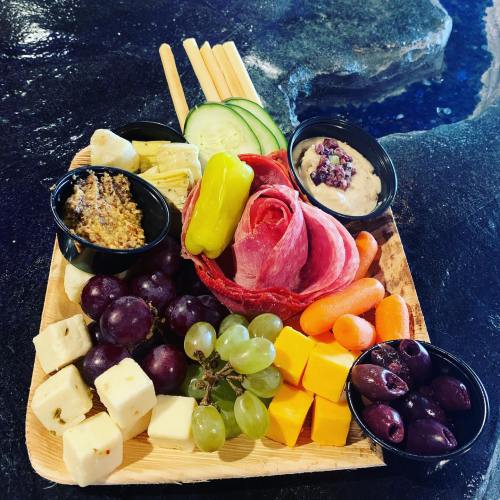 A wooden platter with assorted cheeses, meats, grapes, olives, carrots, cucumbers, and dipping sauces.